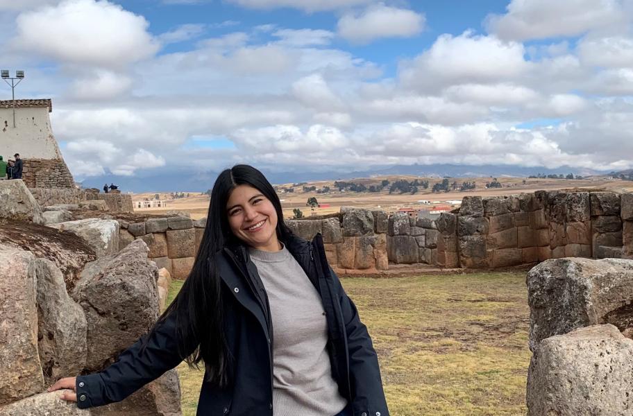 Foto de Carla, mujer sonriente de mediana edad, pelo largo oscuro sobre un fondo de restos arqueológicos de ciudad precolombina